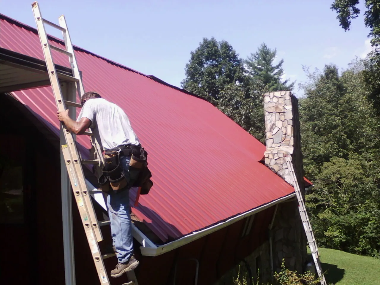 Roofer on a ladder beside a red metal roof and stone chimney — Soffit & Fascia Repair by Roofing Brothers in Noxon